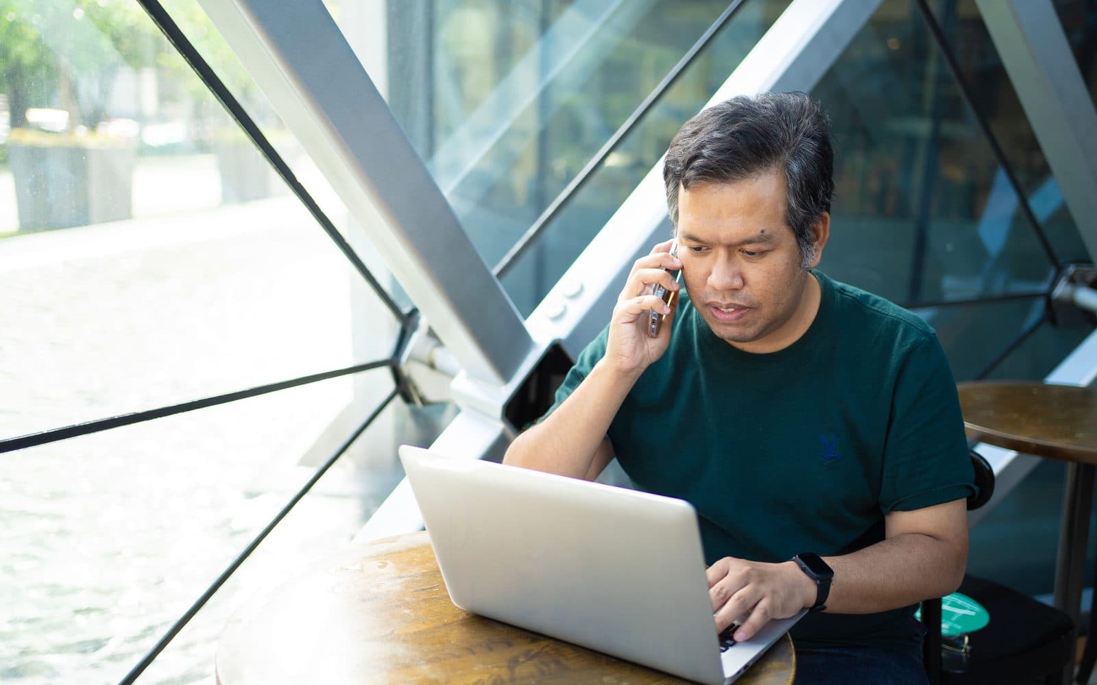 A man taking a phone call while working at a laptop by a window